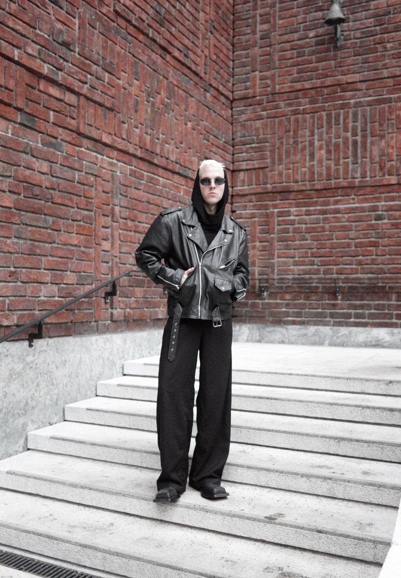 Personne aux cheveux blonds courts portant une veste en cuir noire, un pantalon large noir, des lunettes de soleil et une capuche, debout sur des escaliers en béton à côté d'un mur en briques rouges.