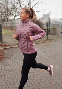Ropa para correr: chaqueta acolchada de color púrpura claro con cuello alto, leggins negros y zapatillas moradas. Entorno al aire libre con pavimento y árboles.