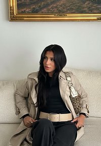 Beżowy trencz na czarnym topie, czarne spodnie, kremowy pasek i torebka w wzory. Długie ciemne włosy; siedzi na teksturowanej sofie.