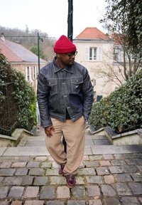 Άνδρας φοράει κόκκινο σκουφί, σκούρο τζιν μπουφάν, μπεζ παντελόνι και καφέ παπούτσια, στέκεται σε πέτρινα σκαλιά που περιβάλλονται από πράσινους θάμνους σε κατοικημένη περιοχή.