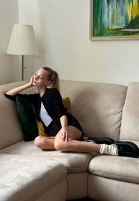 Béžová tkanina gauč s nadýchanými vankúšmi v zelenej a žltej farbe. Osoba v čiernom kardigáne a striebornej blúzke sedí, obutá v čiernych topánkach so bielymi ponožkami.