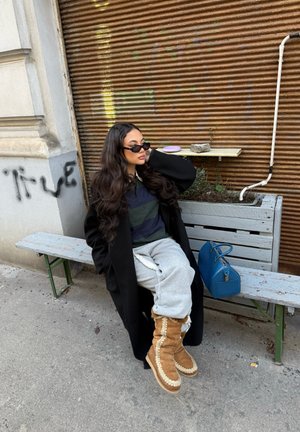 Mujer con el cabello largo, gafas de sol, abrigo negro, pantalones grises de chándal y botas color camel sentada en un banco deteriorado junto a una pared oxidada con un bolso azul.