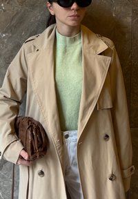 Bež dolg plašč z dvorednim sprednjim delom, svetlo zelena majica, bele kavbojke in rjava pletena torbica. Gladke teksture in večplastni videz.