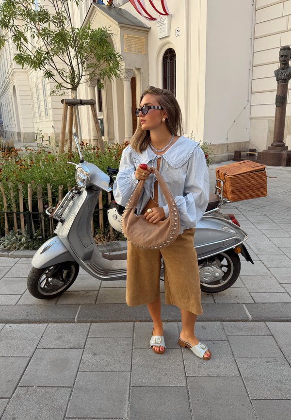 Jeune femme portant des lunettes de soleil, un chemisier blanc et une jupe beige, debout dans la rue, tenant un sac à main beige clouté à côté d'un scooter argenté garé.