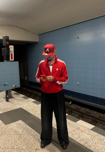 Chaqueta roja de Adidas con rayas blancas, pantalones anchos negros y gorra roja con un logo; persona usando un teléfono en un entorno de estación de metro.