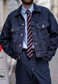 Traper jakna s dvije prednje džepove, srebrnim dugmadi i vidljivim šavovima. Uparena svojim svijetloplavim košuljom i crvenom prugastom kravatom.