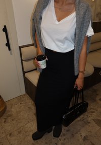 Personne portant un t-shirt blanc, un pull gris posé sur les épaules, une jupe noire, tenant une tasse de café et un sac à main noir à l'intérieur.