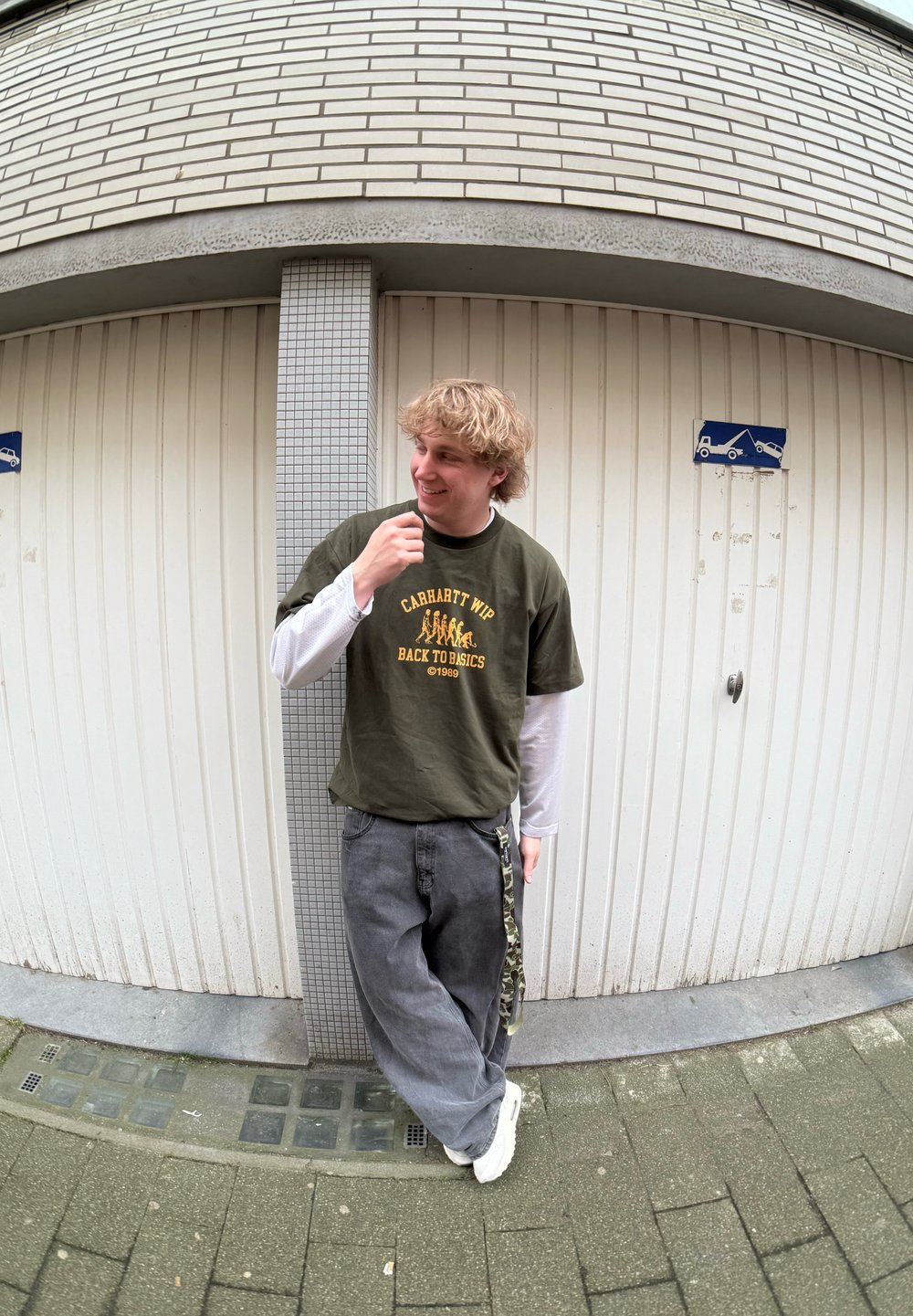 Olivengrøn t-shirt med gul tryk over en hvid langærmet skjorte, grå løstsiddende bukser, hvide sneakers og camouflage detaljer på bukserne.