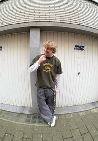 Olivengrøn t-shirt med gul tryk over en hvid langærmet skjorte, grå løstsiddende bukser, hvide sneakers og camouflage detaljer på bukserne.