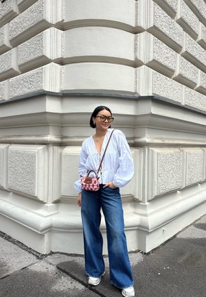 Blusa blanca de manga larga con diseño fruncido, jeans de mezclilla azul de cintura alta y un pequeño bolso de cuadros rosas. Gafas de sol grandes de color amarillo.