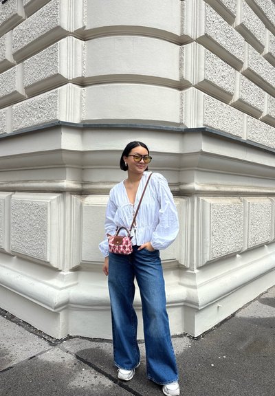 Blusa blanca de manga larga con diseño fruncido, jeans de mezclilla azul de cintura alta y un pequeño bolso de cuadros rosas. Gafas de sol grandes de color amarillo.