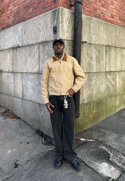 Hombre con chaqueta beige, gorra negra y vaqueros oscuros, de pie junto a la esquina de un edificio de ladrillo y piedra en una calle de la ciudad.