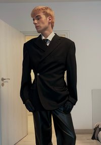 Mustast kohandatud ülikonnajakk ühe nööbiga, kahe esitaskuga, millele on lisatud läikivad mustad nahkseelikud. Kinnastega on välimus täielik.