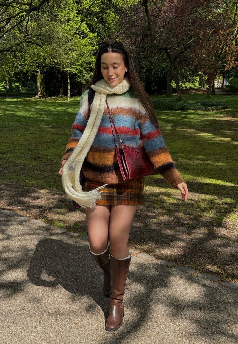Femme marchant dans le parc portant un pull rayé, une jupe à carreaux, une écharpe crème, des bottes marron montant jusqu'aux genoux, et portant un sac rouge au soleil.