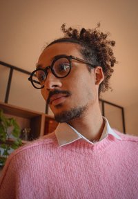 Jeune homme aux cheveux bouclés attachés en chignon, portant des lunettes rondes et un pull rose par-dessus une chemise blanche à col, à l'intérieur avec des étagères en arrière-plan.