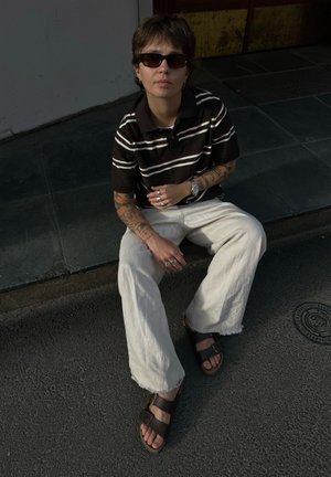 Persona con cabello corto y gafas de sol sentada en la acera, llevando una camisa a rayas, pantalones anchos y claros, sandalias y con los brazos tatuados en un entorno urbano.