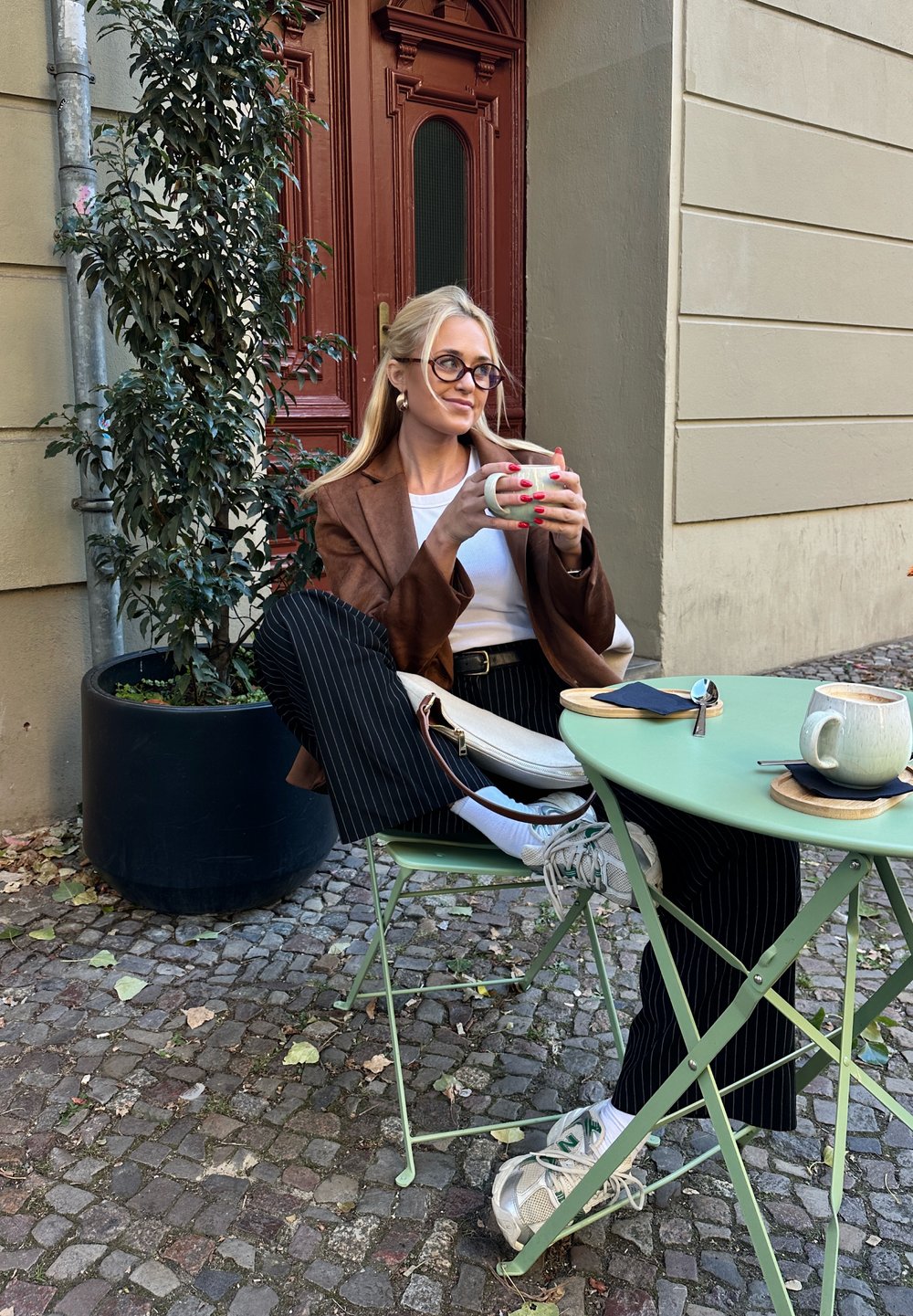 Bruine blazer over een witte top, zwartgestreepte broek en grijze sneakers. Houdt een witte mok vast aan een groene opvouwbare tafel met koffiemokken.