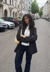 Blazer noir à rayures fines, chemise blanche, jeans bleu foncé et sac à main jaune clair. Rue bordée de voitures garées et bâtiments en arrière-plan.