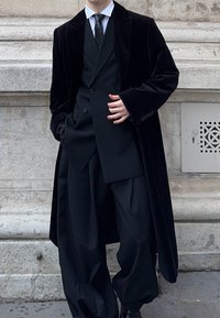 Long manteau en velours noir porté sur un costume trois-pièces noir avec pantalons larges. Chemise blanche et cravate noire complètent le look formel.