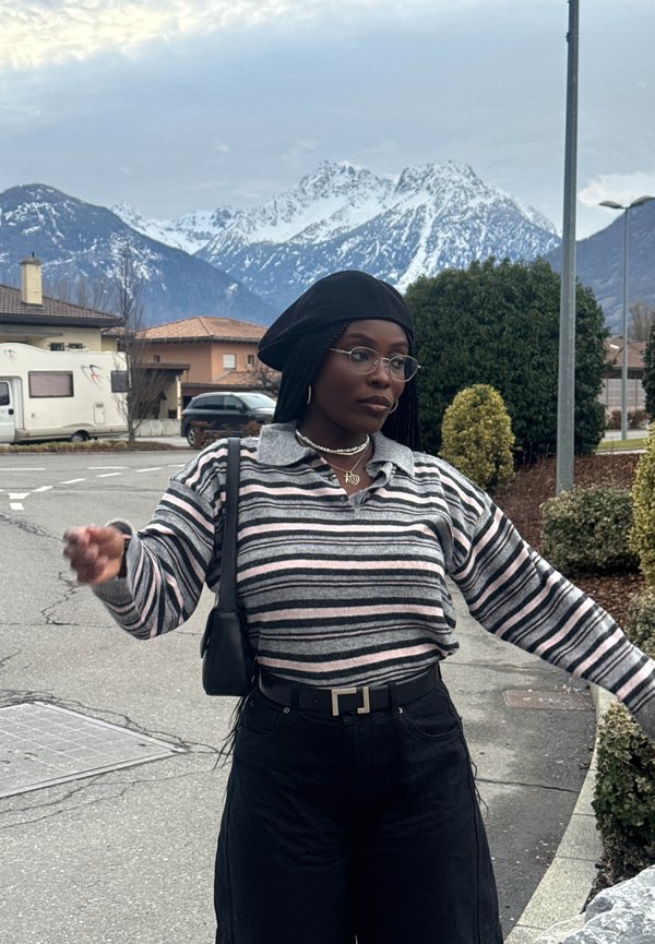 Kvinne i stripete genser og svart beret som poserer i en gate med snødekte fjell og hus i bakgrunnen.