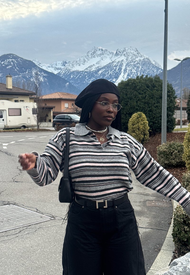 Femme en pull rayé et béret noir posant dans une rue avec des montagnes enneigées et des maisons en arrière-plan.