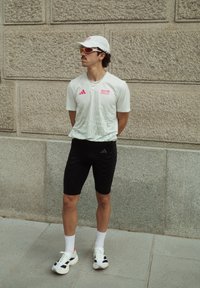 Bărbat îmbrăcat cu un tricou alb de la maratonul BMW Berlin, pantaloni scurți negri, capac alb, ochelari de soare portocalii, stând cu spatele sprijinit de un perete de piatră și cu mâinile la spate.