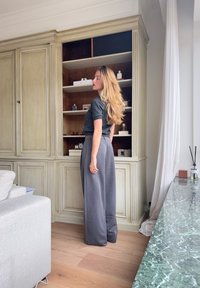 Pantalon gris à jambes larges associé à un haut sombre à manches courtes, le tout dans un intérieur moderne avec des étagères en bois et un comptoir en marbre.