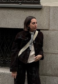 Juoda kordurovinė striukė su blizgiu paviršiumi, derinama su šviesiai smėlio spalvos megztiniu ir tamsiomis kelnėmis. Kaklą puošia šviesiai pilkas šalikas, surištas mazgu.