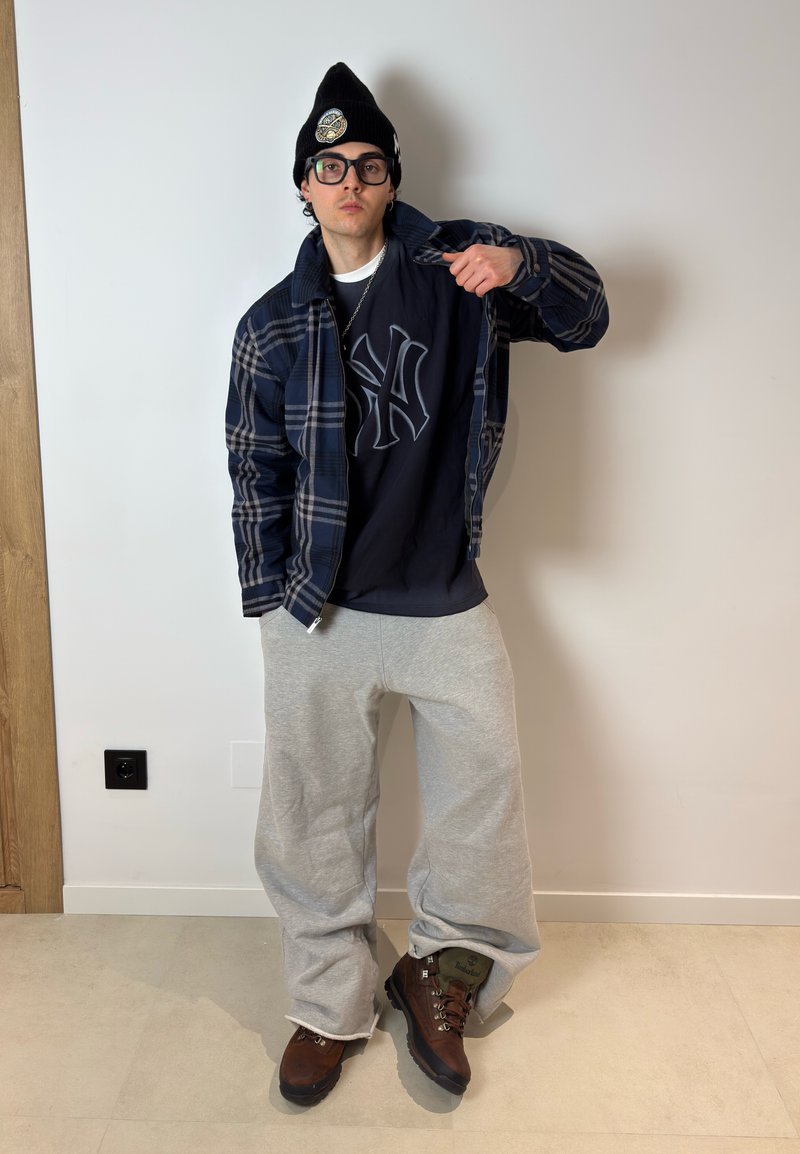 Jeune personne portant un bonnet noir, des lunettes, une veste à carreaux, un t-shirt noir avec un logo, un pantalon de survêtement gris et des bottes marron, debout contre un mur blanc.