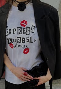 Biała koszulka z czarno-czerwonym tekstem graficznym, odciski czerwonej szminki, w zestawie z czarnym żakietem i czarną torebką w kwiaty. Czerwony pierścionek noszony na ręce.