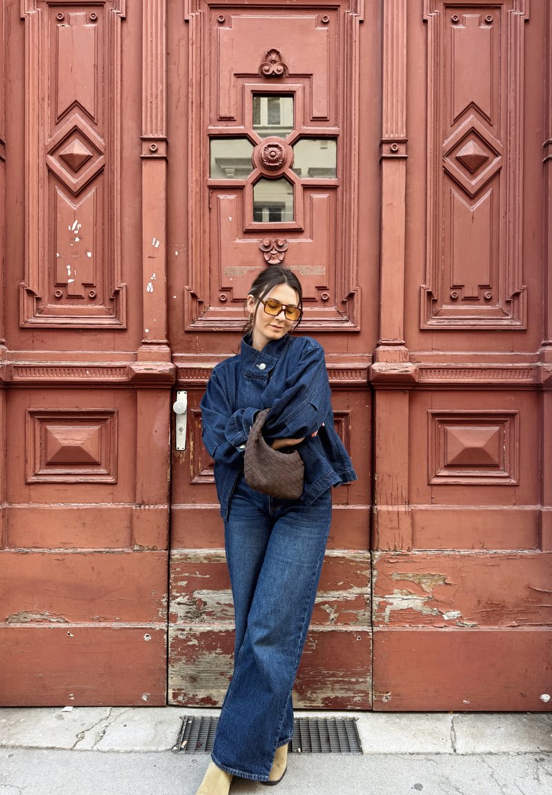 Femme portant une veste en jean oversize et un jean, des bottes beiges et des lunettes de soleil jaunes, posant devant une grande porte en bois rouge ornée, tenant un sac à main tressé marron.