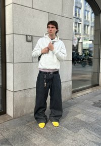 Fehér kapucnis felső, lila póló, laza fekete csíkos nadrág, sárga cipők, kulcsokat tart a kezében, egy textúrált kőfal és üvegablak közelében áll.