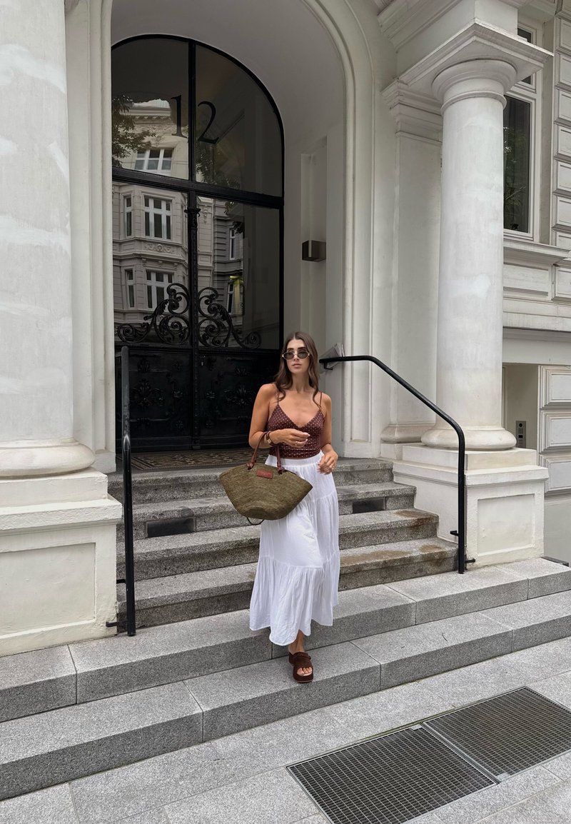 Braun gemusterter Crop-Top, weiße, fließende Maxikleid und Strohtasche. Steht auf Steinstufen in der Nähe eines eleganten Eingangsgebäudes.