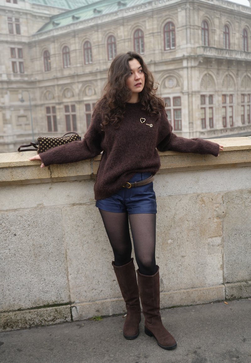 Pull marron duveteux avec une épingle en forme de cœur, short bleu foncé, collants noirs et bottes marron montant jusqu'aux genoux. En arrière-plan, un bâtiment historique.