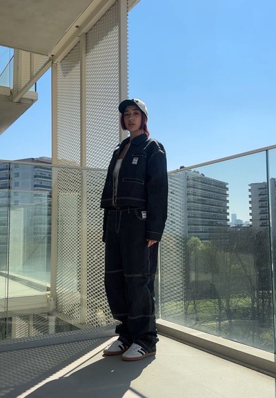 Chaqueta y pantalones de mezclilla a rayas negras con costuras blancas; combinados con zapatillas blancas y una gorra gris, de pie en un balcón.