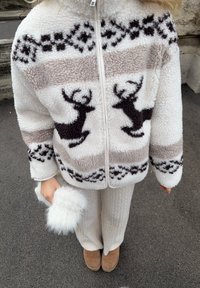 Fliisist lukuga jakk, millel on pruunid põhjapõdrakujud, kreemikas taust ja geomeetrilised detailid. Sobitatud valgete kootud pükste ja pruunide saapadega.