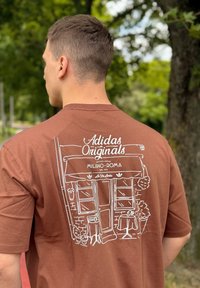 Brązowy bawełniany t-shirt z białym rysunkiem kawiarni, z parasolami, roślinami oraz napisem "Adidas Originals" na plecach.