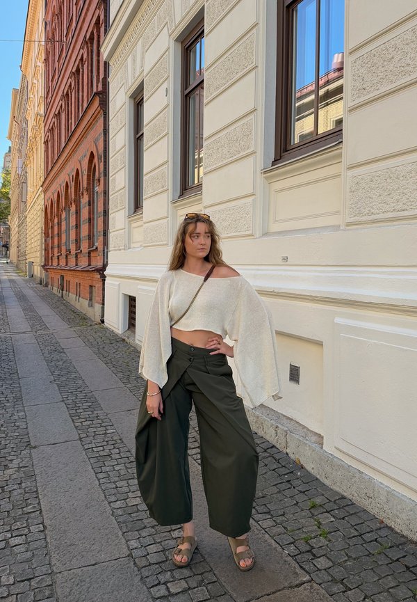 Jeune femme debout dans une rue pavée à côté d'un bâtiment en briques crème et rouge, portant un pantalon large noir, un haut blanc ample et des sandales.