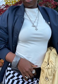 Casaco bomber preto por cima de uma blusa branca justa, pulseira riscada no pulso, colares de prata em camadas com pendentes, e uma bolsa amarela.