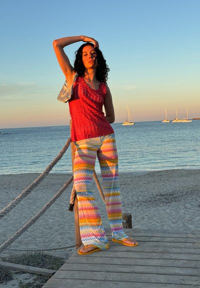 Top de tirantes rojo texturizado, pantalones con patrón de zigzag multicolor, chanclas amarillas; escenario: playa con veleros, iluminación de atardecer.