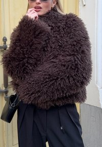 Pull surdimensionné marron, duveteux, en tissu texturé, avec un col montant et des manches larges, assorti à un pantalon noir et un petit sac.