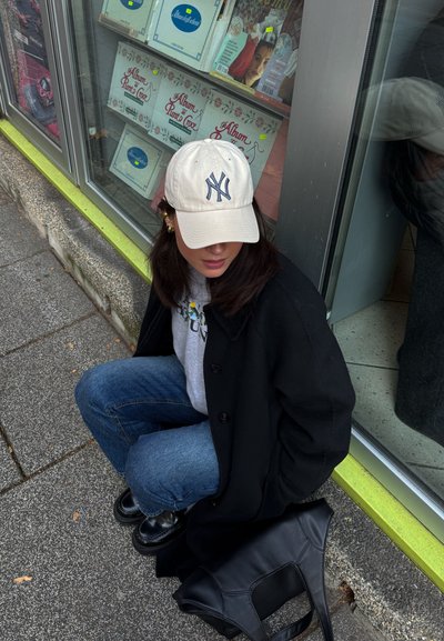 Abrigo negro, sudadera gris, jeans azules y una gorra de béisbol crema con logo azul marino. Zapatos negros y una bolsa negra sobre el pavimento gris.