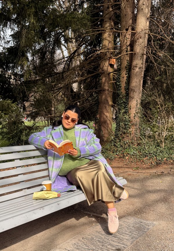 Kobieta w fioletowym płaszczu w kratę i beżowej spódnicy czyta książkę na ławce w parku z kawą i kanapką, otoczona wysokimi drzewami i zielenią.