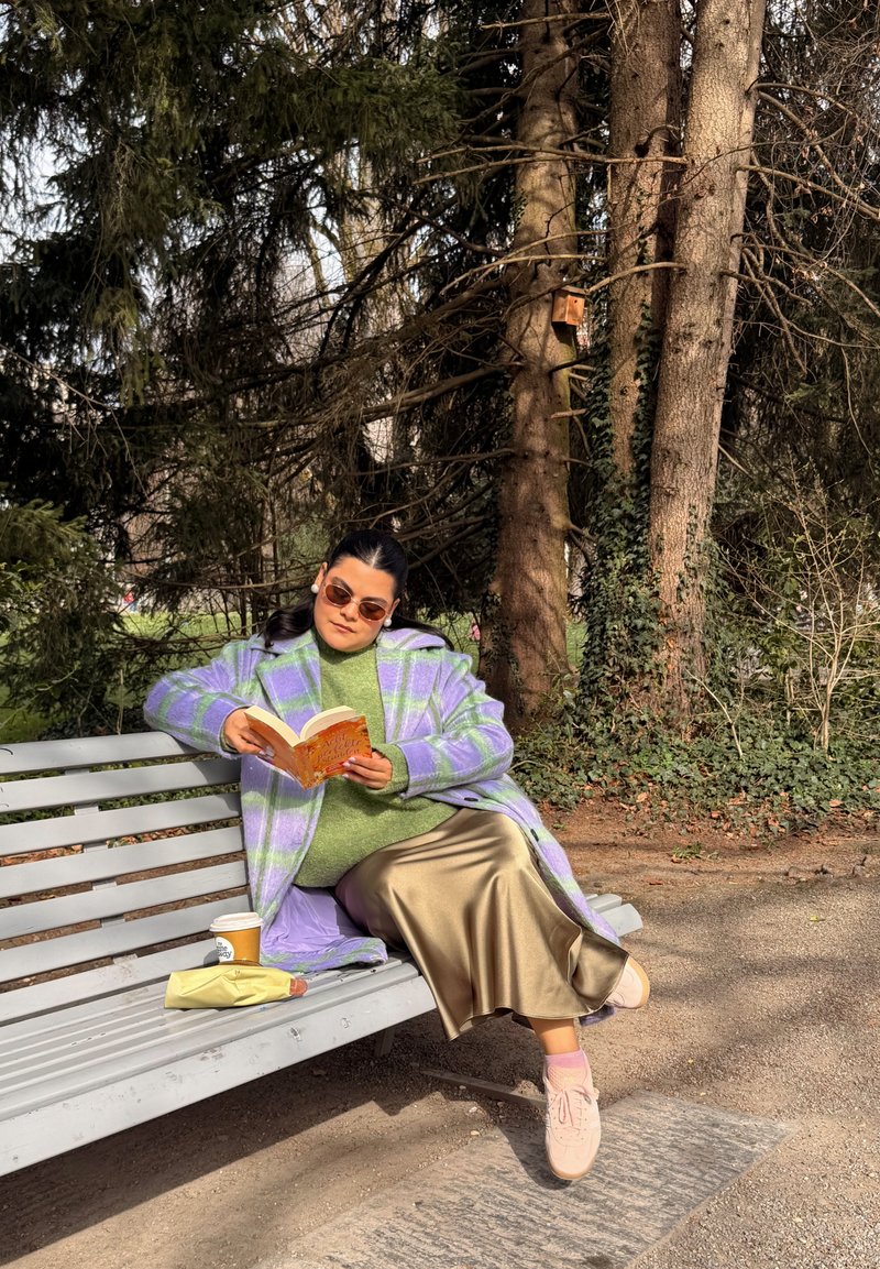 Moteris violetiniu languotu paltu ir smėlio spalvos sijonu skaito knygą ant parko suoliuko su kava ir sumuštiniu, apsupta aukštų medžių ir žalumos.