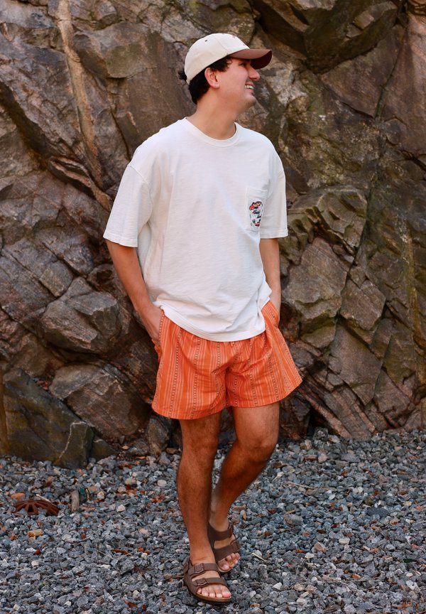 Man wearing orange striped shorts, white t-shirt, white cap, and brown sandals standing on rocky ground by a large stone wall, hands in pockets.