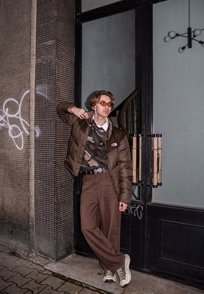 Chaqueta acolchada marrón, suéter con estampado, pantalones marrón oscuro y zapatillas grises. Accesorios incluyen gafas de sol naranjas y un cinturón con estampado.