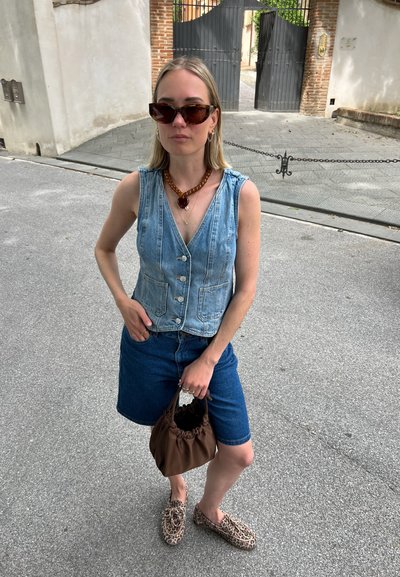 Chaleco de mezclilla sin mangas, azul con botones, combinado con pantalones cortos de mezclilla azul. Bolsa de mano marrón con frunces, zapatos de animal print estilo slip-on, collar de carey.