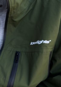 Zielona wodoodporna kurtka z zamkiem błyskawicznym, z czarnym zamkiem i białym logo "lowlights" na przedniej kieszeni. Miękka tekstura.