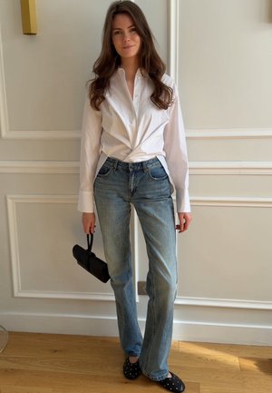 Mujer con el pelo largo y castaño, vistiendo una camisa blanca abotonada y vaqueros azules, sosteniendo un pequeño bolso negro, de pie en el interior.