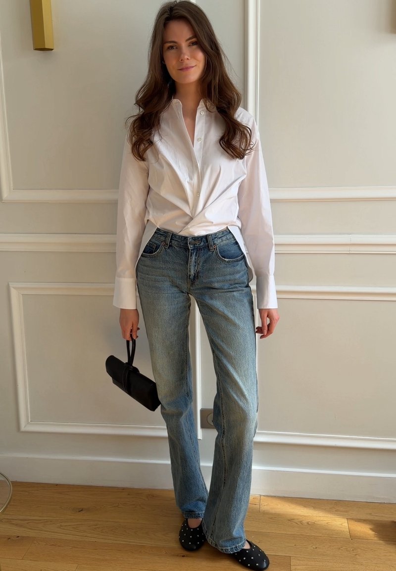 Femme aux longs cheveux bruns portant une chemise blanche boutonnée et un jean bleu, tenant un petit sac à main noir, debout à l'intérieur.
